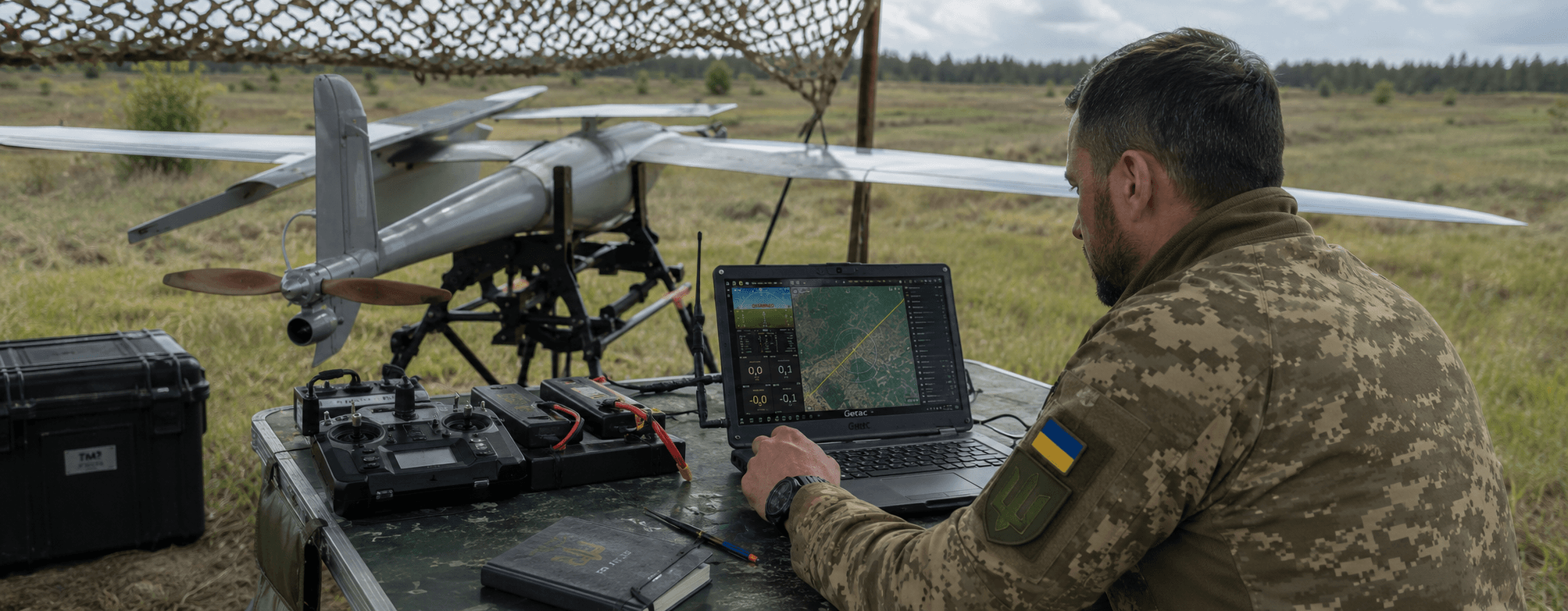 Operator reviewing telemetry at a field laptop next to a drone during a combat test